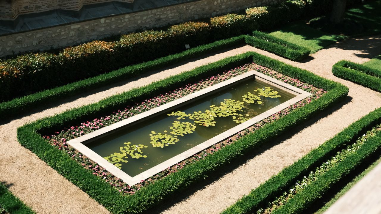 A top view of an elegant rectangular pond surrounded by symmetrical hedges and flowers, in bright daylight.